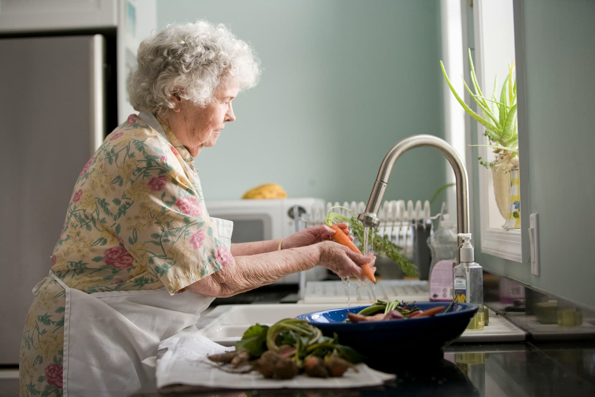 elderly woman in floral dress washing carrots at kitchen sink